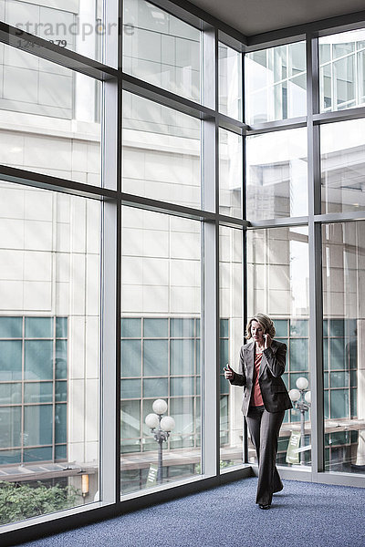 Eine kaukasische Geschäftsfrau  die mit einem Mobiltelefon telefoniert  während sie neben einem großen Fenster in der Lobby eines Kongresszentrums steht.