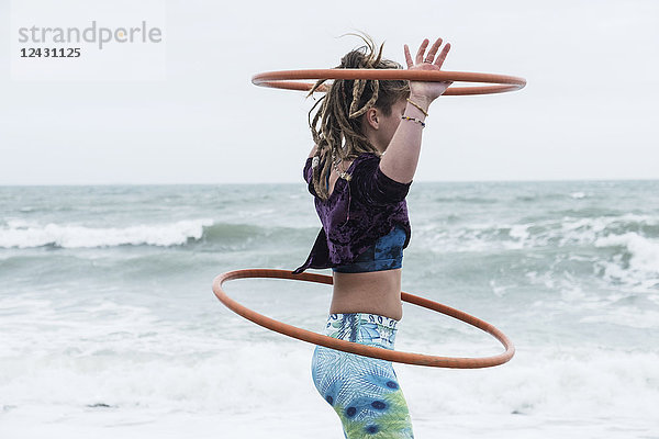 Junge Frau mit braunen Haaren und Dreadlocks  die am Meer steht und zwei Hula-Hoop-Reifen balanciert.