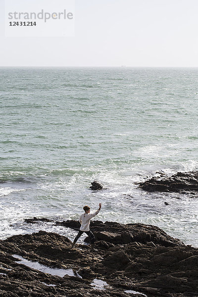 Junge Frau mit braunen Haaren und Dreadlocks in weißer Bluse  die am felsigen Ufer am Meer steht und Tai Chi macht.
