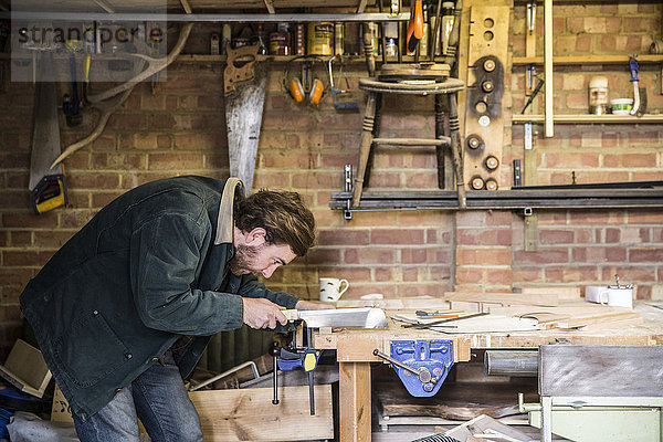 Mann arbeitet stehend an einer Werkbank in einer Holzbearbeitungswerkstatt  mit einer Säge auf einem Stück Holz.