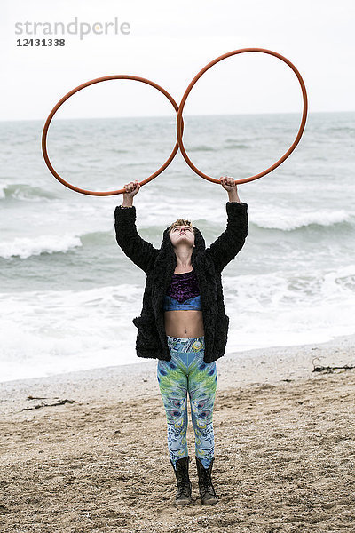 Junge Frau mit braunen Haaren und Dreadlocks  die an einem Sandstrand am Meer steht und zwei Hula-Hoop-Reifen balanciert.