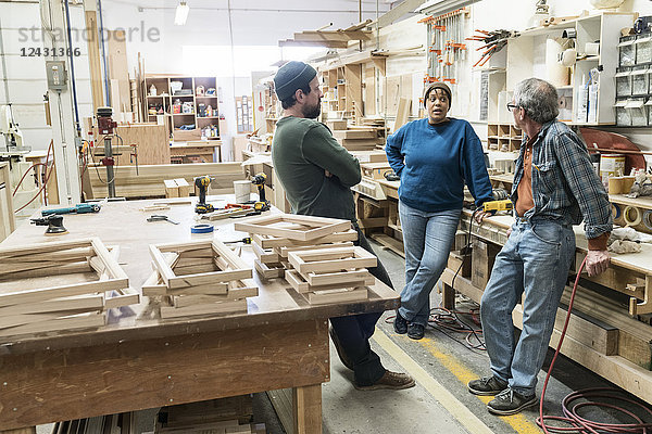 Eine Gruppe gemischtrassiger Tischler  die an einem Arbeitsplatz in einer großen Holzwerkstatt ein Projekt diskutiert.
