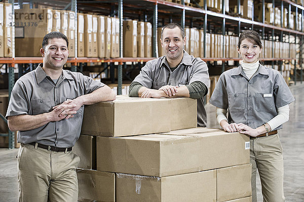 Ein Teamporträt von drei gemischtrassigen uniformierten Lagerarbeitern in einem großen Vertriebslager  umgeben von verpackten Produkten.