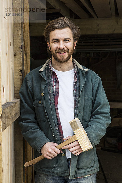 Bärtiger Mann steht in der Türöffnung einer Holzwerkstatt  hält einen Holzschlegel in der Hand und lächelt in die Kamera.