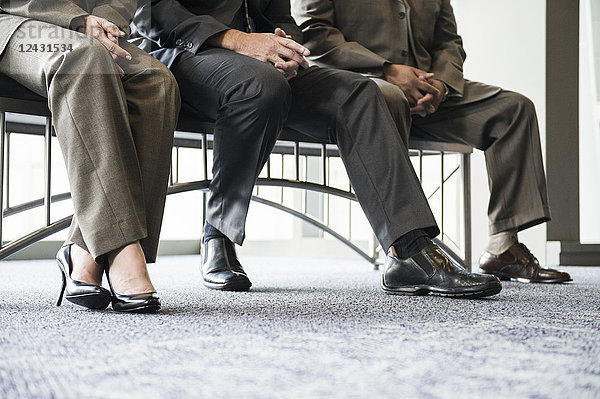Eine Nahaufnahme der Hände und Unterschenkel von drei Geschäftsleuten  die auf einer Bank in einer Bürovorhalle sitzen.