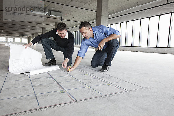 Ein Architekt und ein Geschäftsinhaber kauern über Architekturplänen und zeichnen auf einen Betonboden und planen einen neuen Büroraum in einem leeren  roh gebauten Raum.