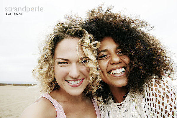 Porträt von zwei lächelnden jungen Frauen mit blonden und braunen Lockenhaaren  die am Sandstrand stehen und in die Kamera schauen.