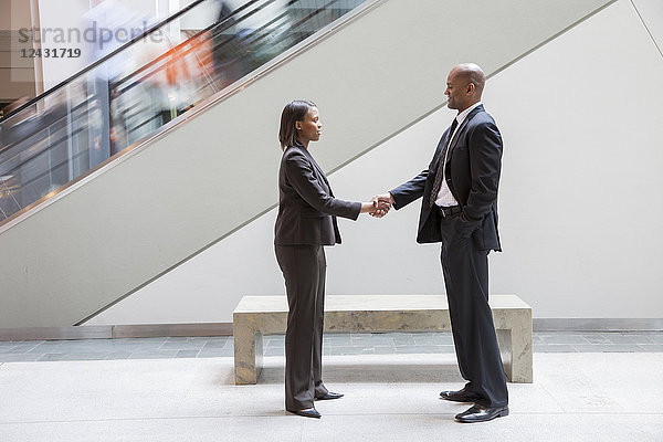Eine schwarze Geschäftsfrau schüttelt einem schwarzen Geschäftsmann  der neben einer Rolltreppe in einem Kongresszentrum steht  die Hand.