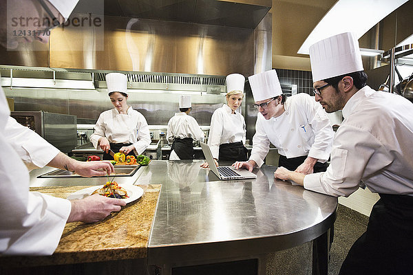 Ein Kochteam arbeitet in einer Großküche  während mehrere Köche an einem Laptop-Computer arbeiten.