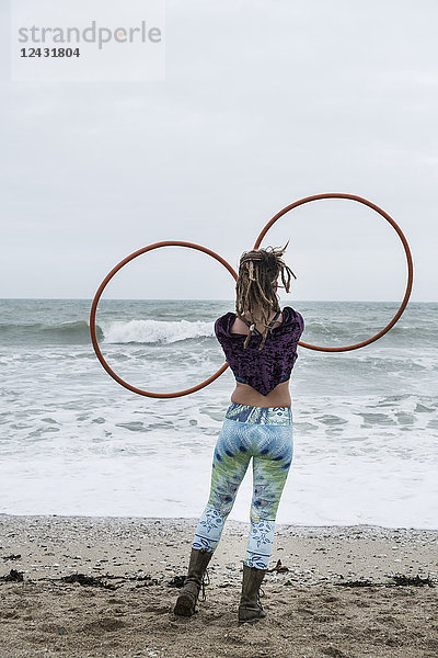 Junge Frau mit braunen Haaren und Dreadlocks  die an einem Sandstrand am Meer steht und zwei Hula-Hoop-Reifen balanciert.