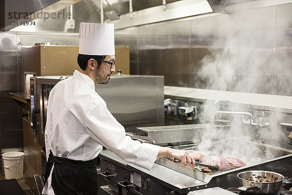 Ein asiatischer Küchenchef  der in einer Großküche mit frischem Fleisch auf dem Grill arbeitet