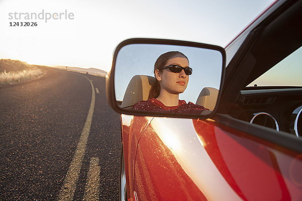 Eine junge Kaukasierin spiegelt sich im Seitenspiegel eines Cabrio-Sportwagens auf der Autobahn.