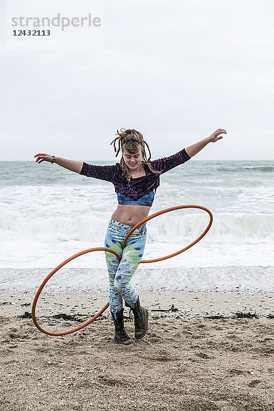 Junge Frau mit braunen Haaren und Dreadlocks  die an einem Sandstrand am Meer steht und zwei Hula-Hoop-Reifen balanciert.