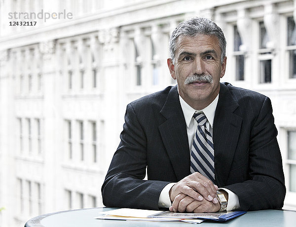 Ein Geschäftsmann aus dem Nahen Osten sitzt an einem Schreibtisch vor einem Fenster  das auf ein Gebäude des Finanzzentrums blickt.