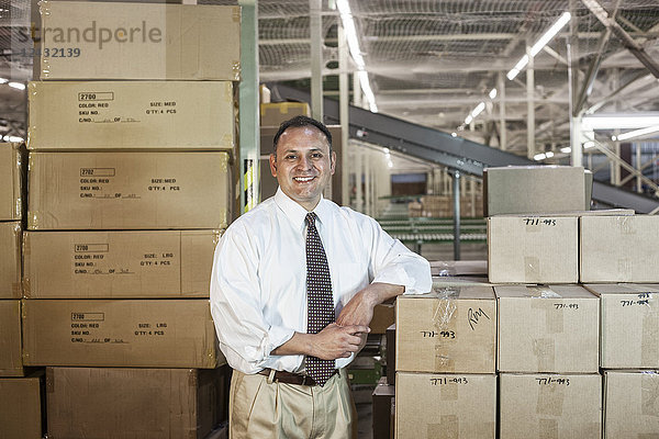 Porträt eines männlichen hispano-amerikanischen Exekutivbeamten in Hemd und Krawatte  umgeben von Produkten  die in Pappkartons in einem großen Vertriebslager gelagert werden.