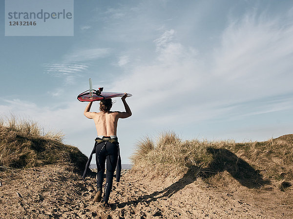 Mann  der mit einem auf dem Kopf balancierenden Surfbrett auf einem Pfad in den Sanddünen Richtung Meer läuft