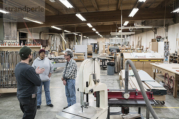 Eine Gruppe gemischtrassiger Tischler  die an einem Arbeitsplatz in einer großen Holzwerkstatt ein Projekt diskutiert.