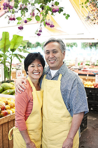 Ein Mann und eine Frau  die einen Obst- und Gemüsemarkt betreiben  tragen Schürzen  stehen nebeneinander und lächeln in die Kamera.