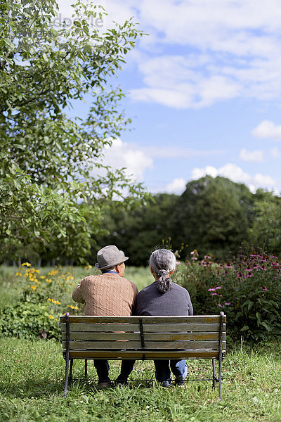 Ehemann und Ehefrau  Rückansicht eines älteren Mannes mit Hut und einer Frau  die Seite an Seite auf einer Bank in einem Garten sitzen.