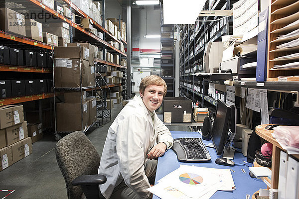Das Porträt eines männlichen kaukasischen Technikers  der an seinem Schreibtisch in einer Ersatzteilabteilung eines technischen Forschungs- und Entwicklungsstandorts sitzt.