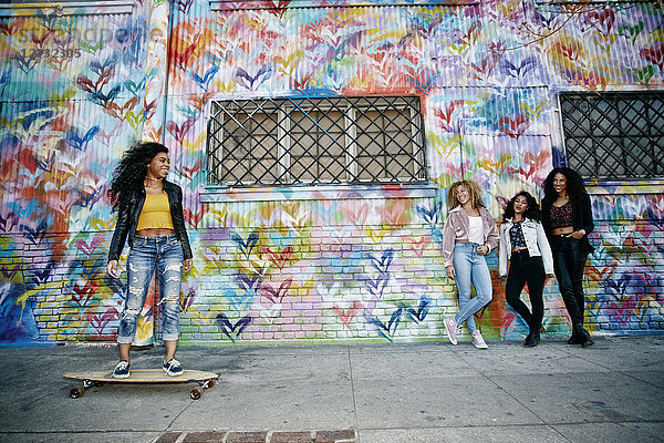 Vier junge Frauen mit lockigem Haar stehen mit bunten Graffiti bedeckt vor dem Fensterladen  eine fährt Skateboard.