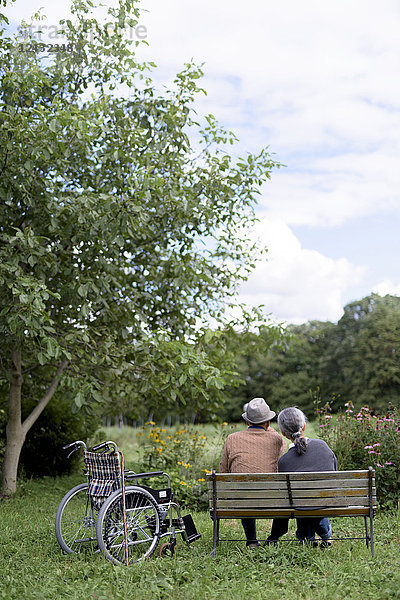 Ehemann und Ehefrau  Rückansicht eines älteren Mannes mit Hut und einer Frau  die Seite an Seite auf einer Bank in einem Garten sitzen  neben der ein Rollstuhl geparkt ist.