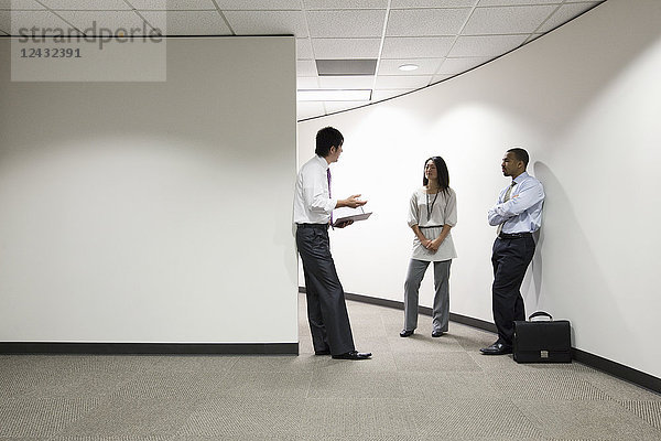 Eine gemischtrassige Gruppe von drei Geschäftsleuten  die in einem Büroflur stehen und reden.