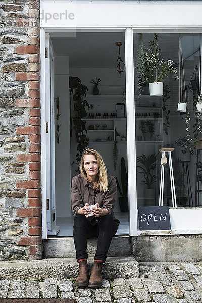 Lächelnde Besitzerin eines Pflanzenladens sitzt auf der Treppe vor ihrem Laden und schaut in die Kamera.