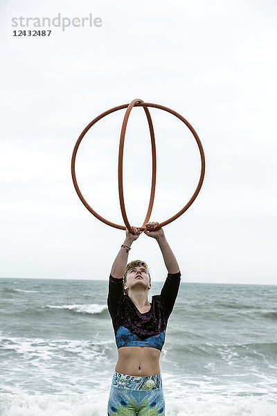 Junge Frau mit braunen Haaren und Dreadlocks  die am Meer steht und zwei Hula-Hoop-Reifen balanciert.