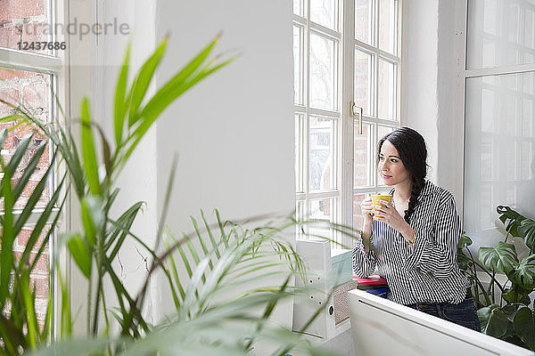 Smiling woman with cup of coffee looking out of window in office