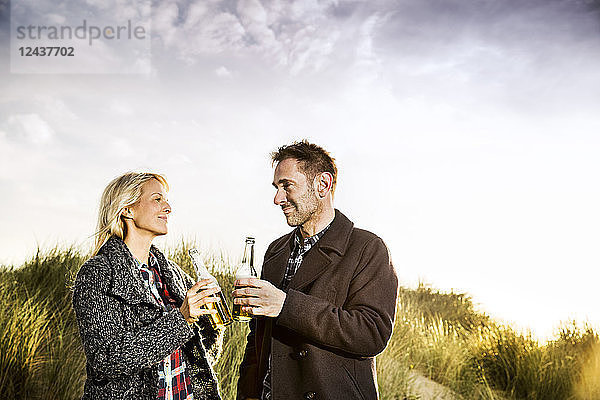 Smiling couple in dunes clinking beer bottles