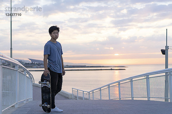 Young Chinese man with skateboard at the beach at sunrise