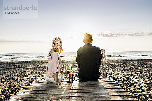 Couple with wine glasses sitting on boardwalk on the beach at sunset