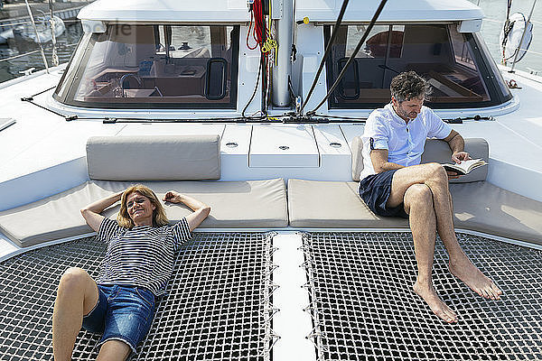 Mature woman relaxing on catamaran trampoline  while husband is reading a book