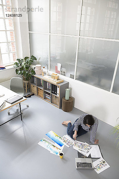 Casual woman with sheets of paper and laptop sitting on the floor in a loft office