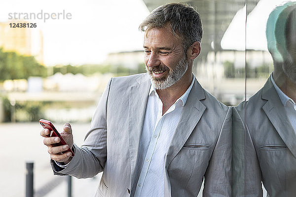 Portrait of smiling businessman leaning against glass facade looking at smartphone