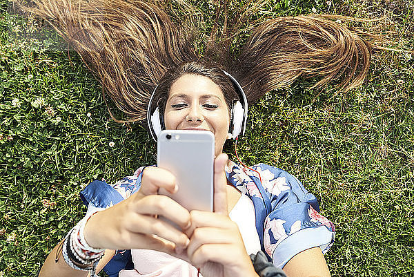 Portrait of smiling woman lying on a meadow listening music with headphones and smartphone
