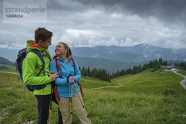 Germany  Lenggries  happy young couple with hiking poles and backpacks on Alpine meadow