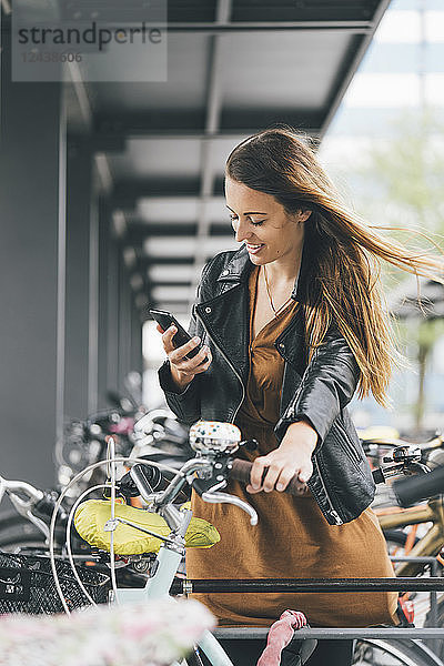 Smiling young woman with bicycle using cell phone in the city