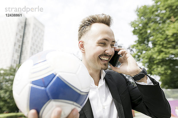 Happy young businessman with football talking on cell phone in the city