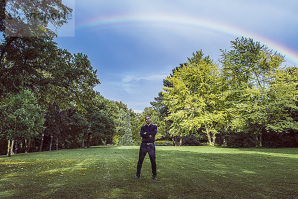 Young businessman standing in park under a rainbow  with arms crossed