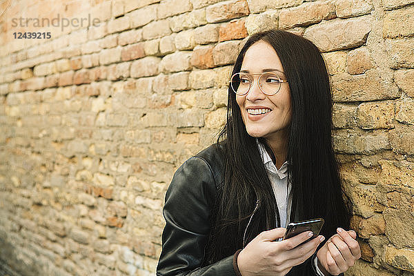Smiling young woman with cell phone at brick wall looking around
