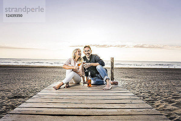 Happy couple with wine glasses sitting on boardwalk on the beach at sunset