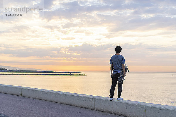Young Chinese man with skateboard standing on wall at the beach