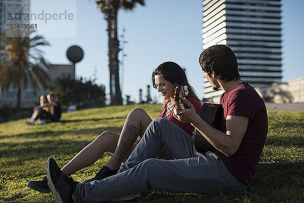 Spain  Barcelona  young man playing guitar for girlfriend sitting on a meadow