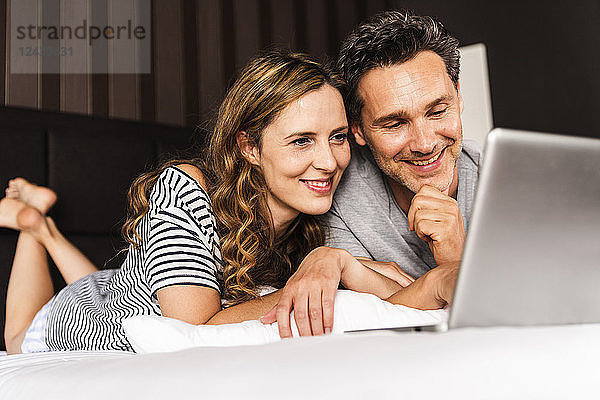 Happy couple lying on bed at home looking at laptop