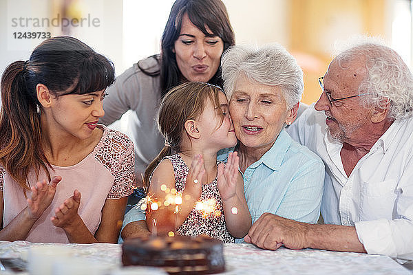 Little girl lwatching sparklers on a birthday cake  sitting on grandmother's lap  with family around