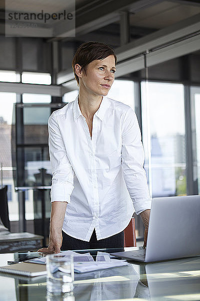 Businesswoman standing at glass table in office with laptop looking sideways