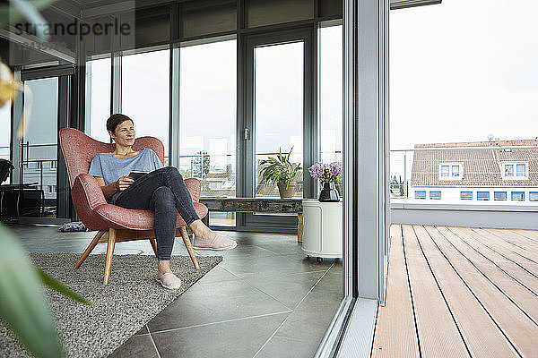 Woman sitting in armchair at home with tablet looking out of balcony door