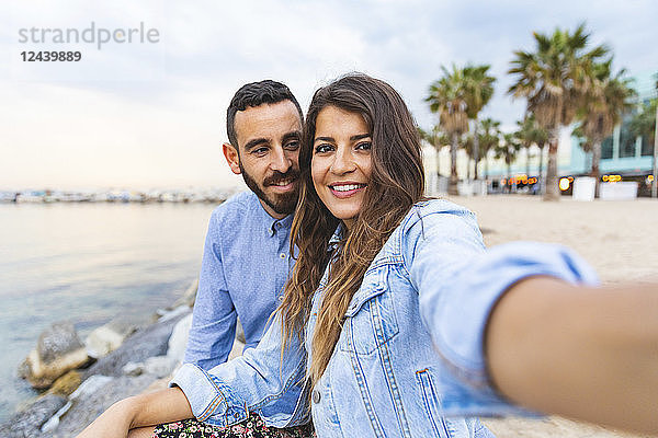 Spain  Barcelona  smiling couple taking a selfie at the seaside